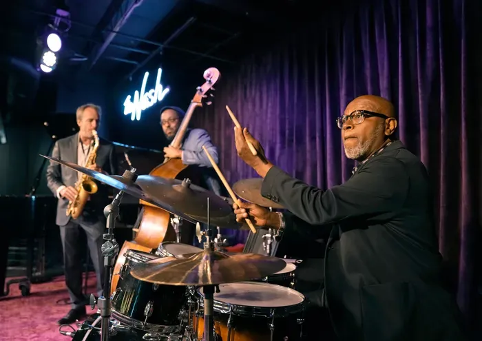 Under cool stage lighting and a glowing neon sign, drummer Lewis Nash, poised mid-beat, leads a jazz trio's intimate live performance alongside Corcoran Holt on upright bass and saxophonist Clark Gibson against a backdrop of deep purple drapes.