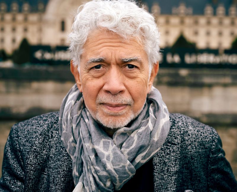 A close-up portrait captures jazz pianist Monty Alexander looking directly forward with a calm, seasoned expression, framed by striking white hair and a softly blurred backdrop of classical architecture.