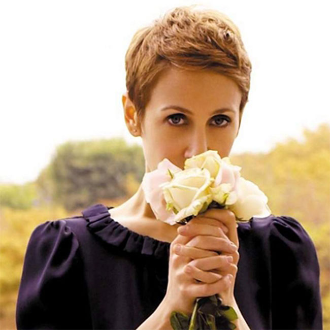 In this intimate close-up, singer Stacey Kent gazes directly forward while holding a delicate bouquet of pale roses close to her face, partially obscuring her nose and mouth. She wears a dark, ruffled blouse that frames her silhouette against a softly blurred, golden-toned outdoor background bathed in warm light.