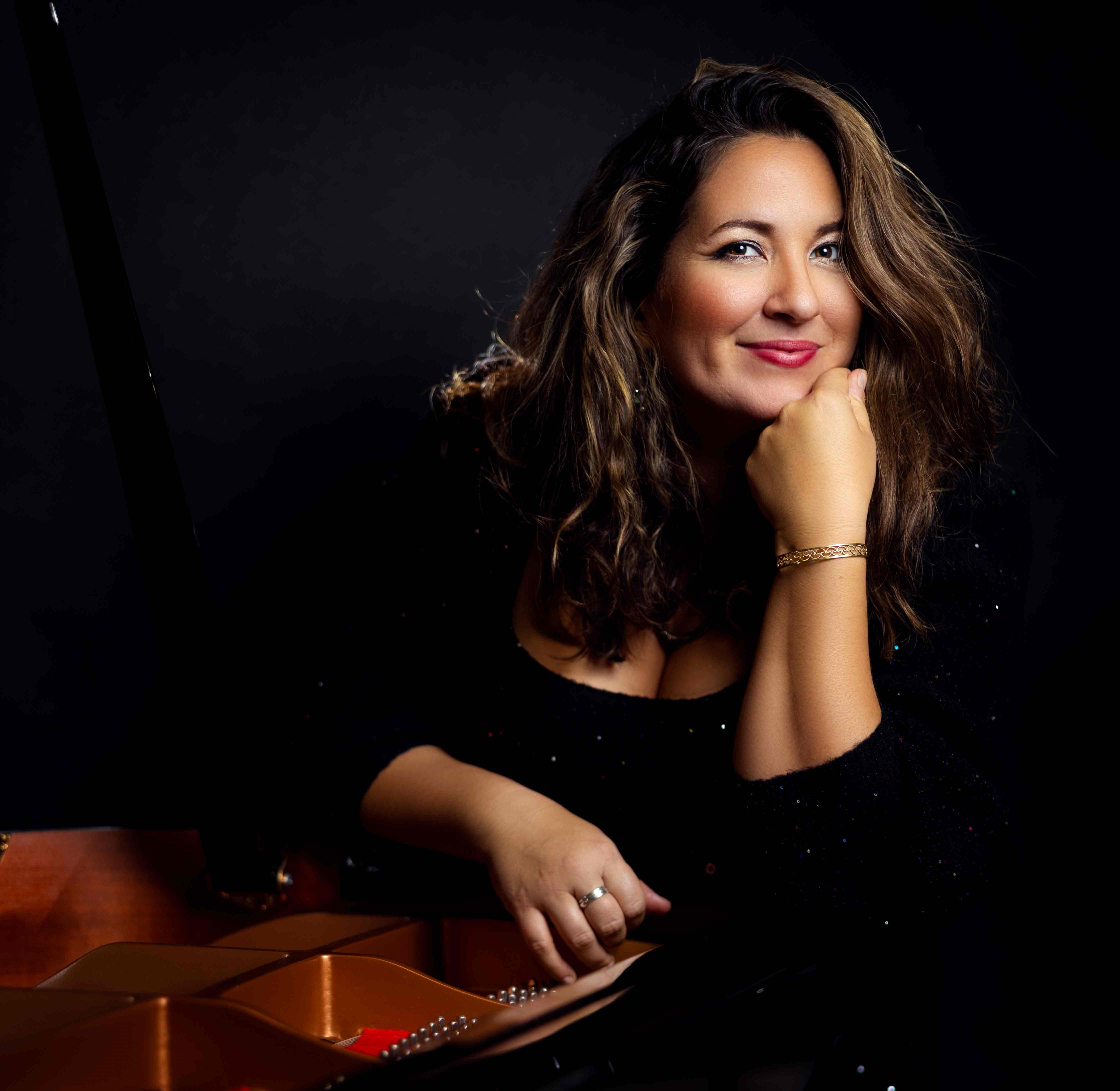 Set against a stark black backdrop, Nicole Zuraitis leans thoughtfully over the gleaming, golden interior of an open grand piano while offering a warm, confident smile.