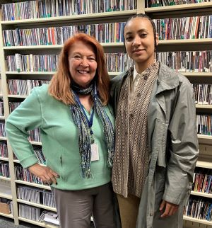 Framed by towering shelves tightly packed with CDs, a warmly smiling red-haired Blaise Lantana in seafoam green stands side-by-side with a composed, stylishly layered esperanza spalding.