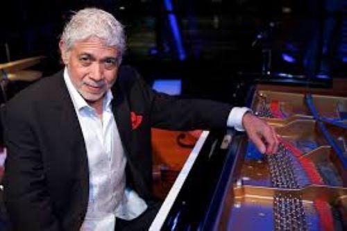 Dressed in a sharp black suit with a pop of a red pocket square, jazz legend Monty Alexander sits at a grand piano, leaning casually against the instrument with a warm, subtle smile on a dimly lit stage.
