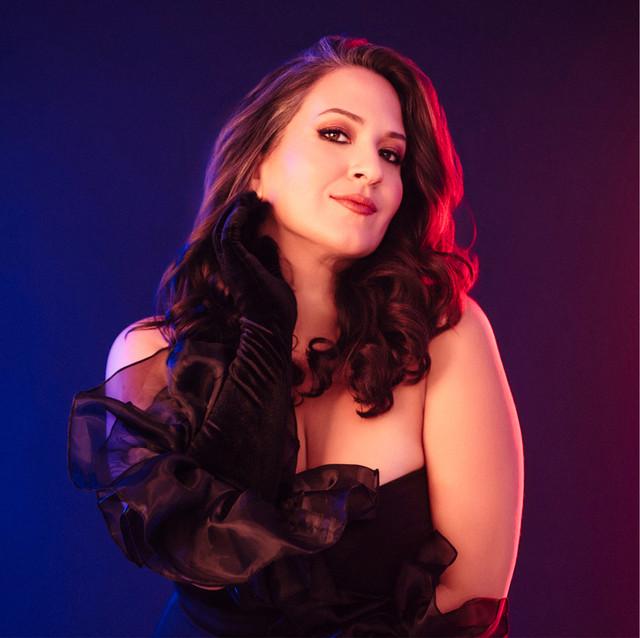 Bathed in a dramatic contrast of cool blue and warm magenta light, Nicole Zuraitis poses with elegant confidence while wearing a strapless black gown and striking, ruffled evening gloves.